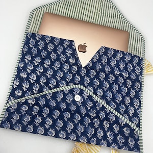 May include: A navy blue laptop sleeve with a floral pattern and a green and white striped interior. The sleeve is open, revealing a gold laptop. A yellow and white striped ribbon is attached to the sleeve.