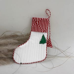 Può includere: Una calza di Natale bianca con polsino e finiture a righe rosse e bianche. Sul davanti è presente un'applicazione di albero di Natale verde. La calza ha un anello a righe rosse e bianche per appenderla.