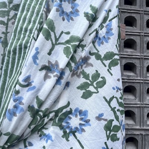 K&ouml;nnte beinhalten: Wei&szlig;er Stoff mit einem floralen Muster in Blau und Grau, akzentuiert durch gr&uuml;ne Bl&auml;tter. Ein gr&uuml;n-wei&szlig; gestreifter Rand ist auf der linken Seite sichtbar. Der Stoff ist drapiert und zeigt das Muster und die Textur.