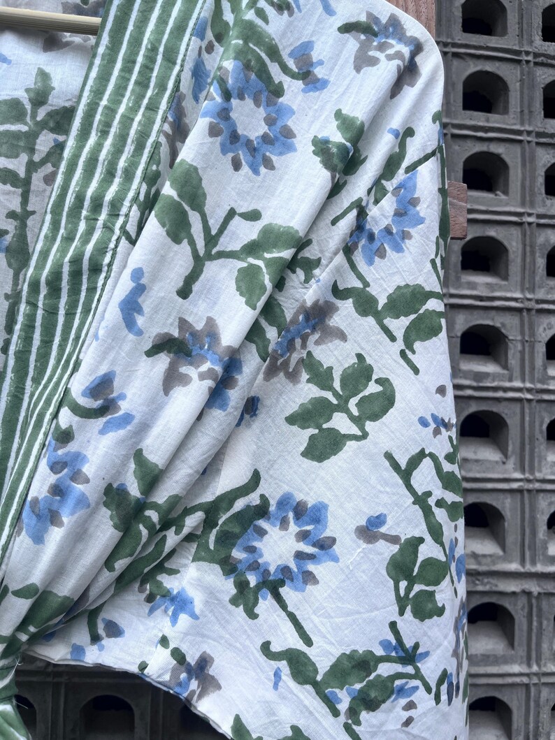 K&ouml;nnte beinhalten: Wei&szlig;er Stoff mit einem floralen Muster in Blau und Grau, akzentuiert durch gr&uuml;ne Bl&auml;tter. Ein gr&uuml;n-wei&szlig; gestreifter Rand ist auf der linken Seite sichtbar. Der Stoff ist drapiert und zeigt das Muster und die Textur.