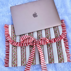 May include: A white laptop sleeve with red and white striped ribbon tied in a bow. The sleeve has a floral pattern and a ruffled edge. A silver laptop is sitting on top of the sleeve.