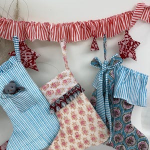 May include: A collection of Christmas stockings in various patterns and colors. The stockings include blue and white stripes, floral prints, and a blue and teal floral design. A red and white striped garland and red star ornaments complete the festive display.