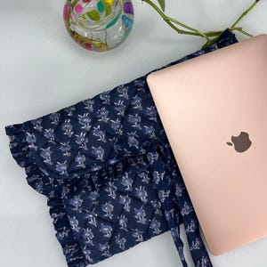 May include: A navy blue quilted laptop sleeve featuring a white floral pattern, a ruffled edge, and tie closures, positioned next to a rose gold laptop. The sleeve is constructed with a diamond-stitched fabric and includes a matching fabric tie.