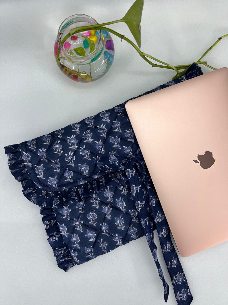 May include: A navy blue quilted laptop sleeve featuring a white floral pattern, a ruffled edge, and tie closures, positioned next to a rose gold laptop. The sleeve is constructed with a diamond-stitched fabric and includes a matching fabric tie.