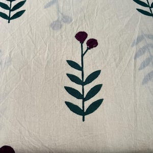 May include: Cream-colored fabric with a repeating pattern of dark green leaves and burgundy flower-like shapes. The fabric has a slightly wrinkled texture, suggesting it may be cotton or linen. The design is simple and botanical.