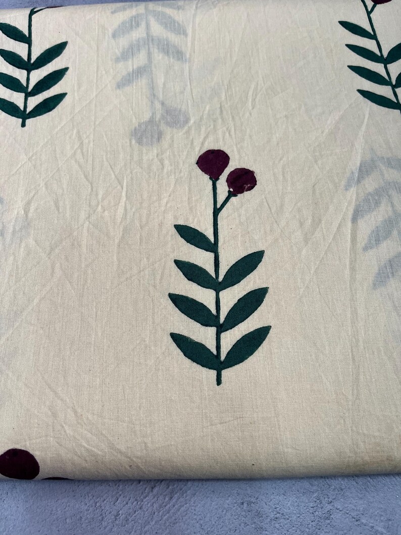 May include: Cream-colored fabric with a repeating pattern of dark green leaves and burgundy flower-like shapes. The fabric has a slightly wrinkled texture, suggesting it may be cotton or linen. The design is simple and botanical.