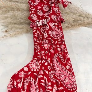 May include: A red Christmas stocking with a paisley and floral pattern in shades of pink and white. The stocking has a quilted texture and a matching fabric bow at the top. The overall design is festive and decorative.