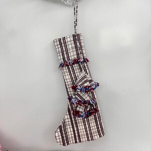 May include: A Christmas stocking with a plaid pattern in white, brown, and burgundy. The stocking is decorated with ruffled accents in red, white, and blue. A matching ribbon is attached for hanging.