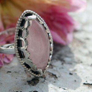 Può includere: Un anello d'argento con una grande pietra ovale di quarzo rosa. La pietra è incastonata in una lunetta decorativa d'argento con un bordo a forma di corda. L'anello è mostrato su uno sfondo sfocato di un fiore rosa.