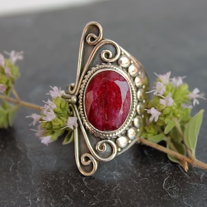 Könnte beinhalten: Ein silberner Ring mit einem großen, ovalen, roten Edelstein, der in eine dekorative Silberfassung gefasst ist.