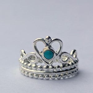 Può includere: Anello a corona color argento con un design a forma di cuore e una piccola pietra turchese al centro. L'anello ha una doppia fascia con una texture a perline. Il design della corona è dettagliato ed elegante.