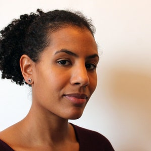 Könnte beinhalten: Eine Frau mit dunkelbraunen, lockigen Haaren, die ein burgunderfarbenes Hemd trägt, blickt direkt in die Kamera. Sie trägt kleine, runde Ohrringe mit einem dunklen Zentrum.