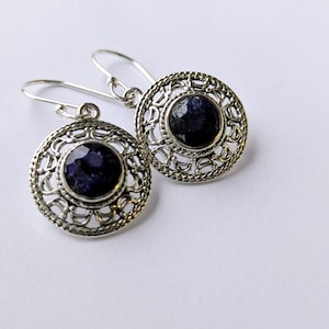 Può includere: Un paio di orecchini color argento con un design circolare. Ogni orecchino presenta una gemma blu scuro sfaccettata incastonata in una cornice traforata ornata. Gli orecchini hanno una chiusura a gancio.