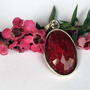 Könnte beinhalten: Ein silberner Anhänger mit einem roten, ovalen Harz-Cabochon. Der Anhänger ist von rosa Blüten umgeben.