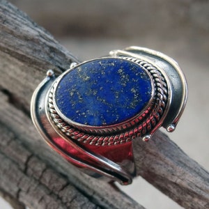 Può includere: Un anello d'argento con una pietra di lapislazzuli blu incastonata in una montatura in argento decorata. L'anello ha un design unico e curvo.