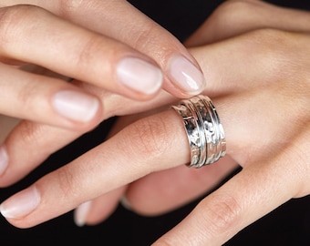 Könnte beinhalten: Ein drehbarer Silberring mit gehämmerter Textur. Der Ring wird an einem Finger getragen und besteht aus mehreren Bändern.