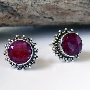 Puede incluir: Un par de pendientes de plata con una piedra preciosa de rubí rojo engastada en cada uno. Los pendientes tienen un diseño circular con un borde texturizado.