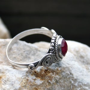 Könnte beinhalten: Ein silberner Ring mit einem roten Edelstein, der in einer dekorativen silbernen Fassung sitzt. Der Ring hat ein gedrehtes Band mit einem Spiralmuster.
