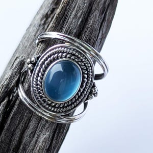 Può includere: Un anello d'argento con una grande pietra preziosa blu ovale. La pietra preziosa è incastonata in una lunetta dettagliata a forma di corda. La fascia dell'anello ha un design unico a vortice. L'anello è esposto su un pezzo di legno stagionato.