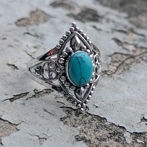 Può includere: Un anello d'argento con una pietra turchese ovale incastonata in un elaborato design a filigrana. L'anello presenta un'incastonatura a forma di diamante attorno alla pietra, con piccoli dettagli di perline. L'anello è esposto su una superficie chiara e strutturata.