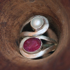 Könnte beinhalten: Ein silberner Ring mit einem roten Edelstein und einer weißen Perle. Der Ring ist in einem Wirbeldesign gefasst.