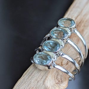 Può includere: Una collezione di anelli in argento, ognuno con una pietra preziosa ovale azzurra. Gli anelli sono esposti su un pezzo di legno chiaro contro uno sfondo scuro. Le pietre preziose sono traslucide.
