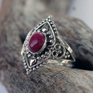 May include: A silver ring with an oval, deep red gemstone at the center. The ring has an ornate design with beaded and swirling details. The ring is set on a piece of weathered wood.