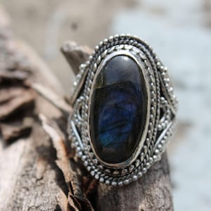 Può includere: Anello d'argento ovale con una grande pietra preziosa blu scuro. L'anello presenta un design dettagliato e strutturato attorno alla pietra e alla fascia. La gemma mostra un gioco di colori, con sfumature di blu e verde.
