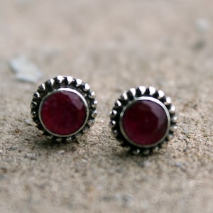 Peut inclure: Une paire de boucles d'oreilles en argent avec une pierre précieuse rouge au centre de chaque boucle d'oreille. Les boucles d'oreilles ont une bordure décorative autour de la pierre précieuse.