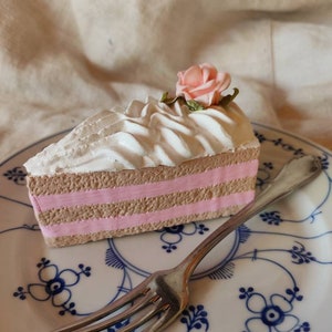 Könnte beinhalten: Ein Stück Kuchen mit rosa Zuckerguss und einer rosa Rose darauf. Der Kuchen liegt auf einem blau-weiß gemusterten Teller mit einer silbernen Gabel.