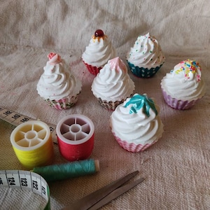 Könnte beinhalten: Sechs Mini-Cupcakes mit weißem Frosting und verschiedenen bunten Streuseln. Die Cupcakes sind auf einer weißen Leinenoberfläche mit Nähzubehör angeordnet, darunter Garnrollen, eine Schere und ein Maßband.