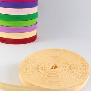 Puede incluir: Una pila de ribetes de tela de colores, incluyendo morado, rosa, naranja, verde y amarillo. Un solo rollo de ribete de tela amarillo se muestra en primer plano.