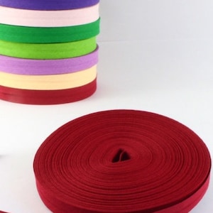 Puede incluir: Una pila de rollos de cinta al bies de colores, incluyendo rojo, morado, verde y naranja. Un rollo separado de cinta al bies roja está desenrollado. La dirección del sitio web jarkashop.de es visible.