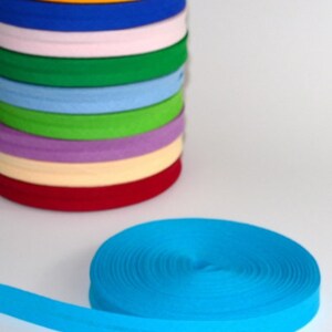 Puede incluir: Una pila de rollos de cinta al bies de colores, incluyendo rojo, amarillo, azul y verde. Un rollo separado de cinta al bies turquesa está desenrollado sobre una superficie blanca. El texto "jarkashop.de" es visible.