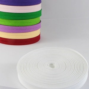 Puede incluir: Una cinta de algodón blanco en twill, enrollada y lista para ser utilizada en proyectos de costura o manualidades. La cinta tiene 25 mm de ancho y se muestra junto a una pila de cintas de algodón en twill de varios colores.