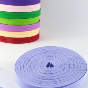 Puede incluir: Una pila de rollos de cinta al bies de colores, incluyendo morado, naranja, rosa y verde. Un rollo separado de cinta al bies morada clara está desenrollado. El sitio web "jarkashop.de" es visible en la esquina inferior derecha.