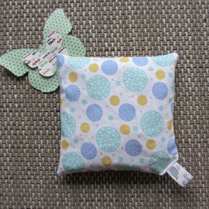 Könnte beinhalten: Ein weißes Kissen mit einem blauen, gelben und grünen Tupfenmuster. Das Kissen liegt auf einer braunen gewebten Oberfläche mit einem grünen Schmetterlingsausschnitt auf der linken Seite.