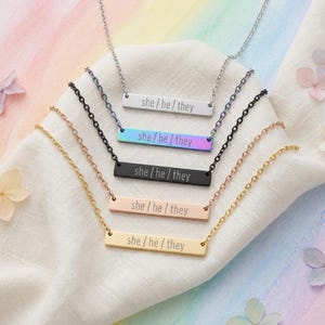 以下が含まれることがあります： she / he / theyという文字が刻印された、5本の水平バーネックレスとチェーンのセット。シルバー、虹色のイリデセント、マットブラック、ローズゴールド、ゴールドの5色で、扇状に並べられています。
