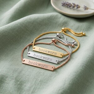 May include: Three adjustable slider bracelets featuring rectangular metal bars in gold, silver, and rose gold tones. Each bar is engraved with the word "bookworm" in a typewriter-style font. The bracelets have delicate box chains.
