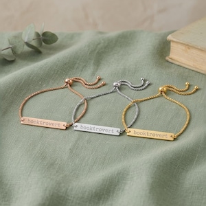 May include: Three adjustable slider bracelets with thin box chains in rose gold, silver, and gold tones. Each bracelet features a rectangular metal bar engraved with the word booktrovert in a simple, lowercase sans-serif font.