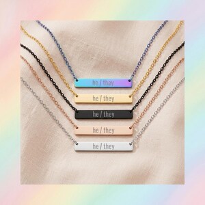 May include: Five horizontal bar necklaces with matching chains, each engraved with the text he / they. The pendants come in iridescent, gold, black, rose gold, and silver finishes, arranged vertically from top to bottom on a light fabric.