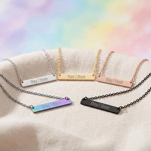May include: Five metal bar necklaces with delicate chains, each engraved with the text they / them. The collection includes silver, gold, rose gold, iridescent rainbow, and matte black finishes, all arranged together in a circular display.