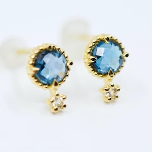 Könnte beinhalten: Ein Paar goldfarbene Ohrstecker. Jeder Ohrring hat einen runden, facettierten, hellblauen Edelstein, der von einer dekorativen Goldfassung umgeben ist. Ein kleiner, sternförmiger Akzent mit einem klaren Stein hängt darunter.