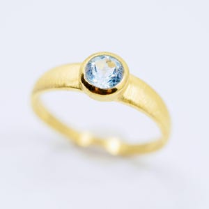 May include: A gold ring with a round, light blue gemstone set in a bezel. The ring has a simple, elegant design with a brushed finish on the band. The gemstone is the focal point, catching the light and adding a touch of color.