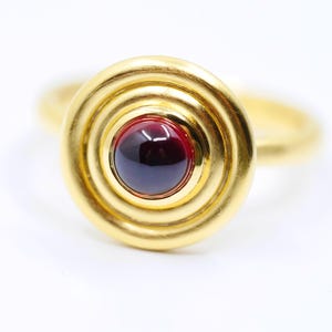 May include: A gold ring with a circular design. The ring features a deep red, circular stone set in the center, surrounded by concentric gold bands. The band of the ring is also gold.