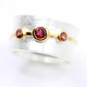 Può includere: Un anello d'argento con tre pietre preziose rosse incastonate in castoni d'oro. L'anello ha una fascia strutturata e una fascia dorata che attraversa il centro, creando un contrasto sorprendente. Le pietre sono equidistanti.