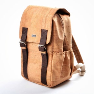 May include: A tan cork backpack with brown leather straps and buckles. The backpack has a zipped pocket on the front and two side pockets.