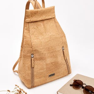 Könnte beinhalten: Ein hellbrauner Kork-Rucksack mit zwei Reißverschlusstaschen auf der Vorderseite. Der Rucksack hat zwei verstellbare Schultergurte und einen oberen Griff. Eine braune Sonnenbrille und ein Notizbuch liegen neben dem Rucksack.