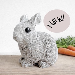 Può includere: Una statuetta di coniglio grigio con occhi neri e pelliccia strutturata è su una superficie di legno. La parola "NEW!" è in un cerchio rosa. Carote visibili sullo sfondo. Questo oggetto decorativo è per l'arredamento della casa.