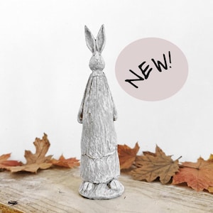 May include: A tall, slender, light gray rabbit figurine with a textured, weathered finish. The rabbit stands upright with long, pointed ears and a smooth, draped body. A circular pink graphic to the right contains the black text NEW!.